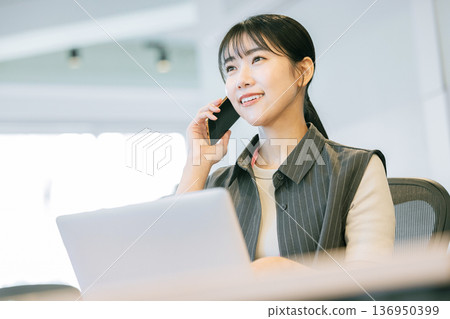 辦公室裡，一名女子一邊用筆記型電腦一邊打電話。 136950399