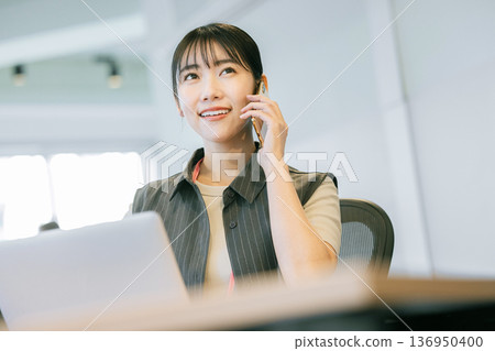 辦公室裡,一名女子一邊用筆記型電腦一邊打電話。 辦公室裡,一名女子一邊用筆記型電腦一邊打電話。 136950400