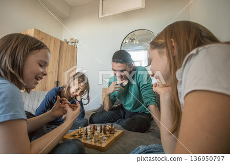 Four people sit on a bed, focused on a chess game. Two adults and two children study the board. 136950797