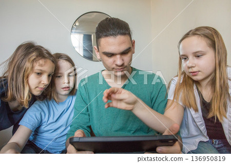 Four children sit together on a couch in a living room. They look at a tablet closely Four children sit together on a couch in a living room. They look at a tablet closely 136950819