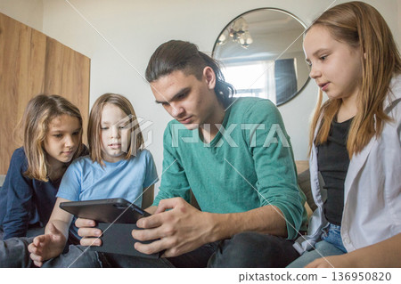 Four children sit together on a couch in a living room. They look at a tablet closely 136950820