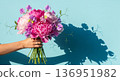A hand holds a beautiful, elegant bouquet of fresh flowers against a bright, light blue background. The arrangement features pink peonies, sweet peas 136951982
