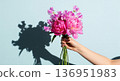 A hand holds a beautiful, elegant bouquet of fresh flowers against a bright, light blue background. The arrangement features pink peonies, sweet peas 136951983