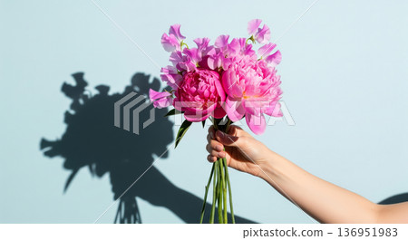 A hand holds a beautiful, elegant bouquet of fresh flowers against a bright, light blue background. The arrangement features pink peonies, sweet peas 136951983