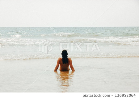 Young woman sitting in shallow ocean water on a sandy beach, view from behind. Moody vintage film style with analog grain. Minimalist coastal lifestyle concept for mindfulness, zen, and summer peace. Young woman sitting in shallow ocean water on a sandy beach, view from behind. Moody vintage film style with analog grain. Minimalist coastal lifestyle concept for mindfulness, zen, and summer peace. 136952064