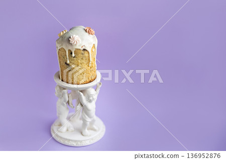Traditional Easter cake with icing displayed on decorative white ceramic stand with angel details, soft lavender background creating elegant festive atmosphere Traditional Easter cake with icing displayed on decorative white ceramic stand with angel details, soft lavender background creating elegant festive atmosphere 136952876
