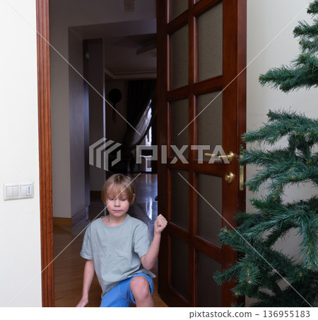 A young Caucasian boy with blonde hair is kneeling on the floor near a wooden door with glass panels. A young Caucasian boy with blonde hair is kneeling on the floor near a wooden door with glass panels. 136955183