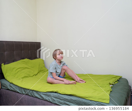 A young Caucasian boy with blonde hair sits barefoot on a bed with a bright green duvet. A young Caucasian boy with blonde hair sits barefoot on a bed with a bright green duvet. 136955184
