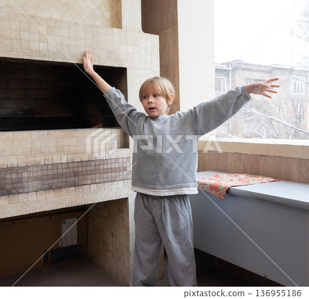 A young Caucasian boy with blonde hair stands on a tiled balcony or porch during winter. He is wearing a grey sweatshirt and sweatpants. A young Caucasian boy with blonde hair stands on a tiled balcony or porch during winter. He is wearing a grey sweatshirt and sweatpants. 136955186