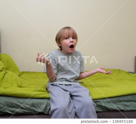 A young Caucasian boy with blonde hair sits on the edge of a bed with bright green bedding. He is wearing a grey t-shirt and sweatpants. 136955188