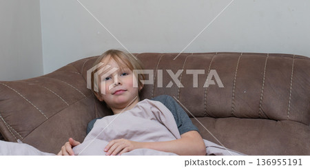 A portrait of a young Caucasian boy with blonde hair resting on a brown sofa. A portrait of a young Caucasian boy with blonde hair resting on a brown sofa. 136955191