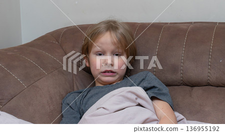 A close-up, eye-level shot of a young Caucasian boy lying on a brown sofa, covered by a light beige blanket. A close-up, eye-level shot of a young Caucasian boy lying on a brown sofa, covered by a light beige blanket. 136955192