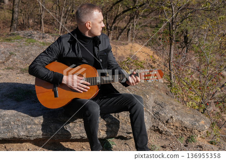 Portrait of smiling caucasian artist plays wooden acoustic guitar in autumnal park. Young songwriter plays string musical instrument hobby outside in nature fall time. Audio music healing Portrait of smiling caucasian artist plays wooden acoustic guitar in autumnal park. Young songwriter plays string musical instrument hobby outside in nature fall time. Audio music healing 136955358