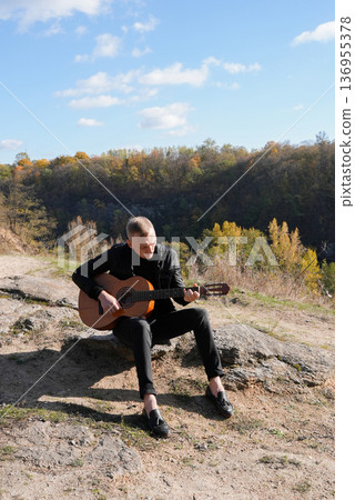 Blonde man playing string guitar outdoors in autumn forest. Concept of sound therapy, mental health and wellness rituals. Calmness tranquility audio-sensory practices. Aura farming energy 136955378