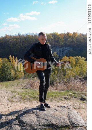 Beautiful blonde man playing acoustic guitar and singing relaxing enjoying outdoors on sunny autumn day. Artist learning and practicing playing musical instrument. Fall leaves autumnal 136955396