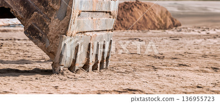 The excavator's bucket digs into fine sand, revealing the energy of construction work at a coastal site, where progress meets natural beauty and earth 136955723