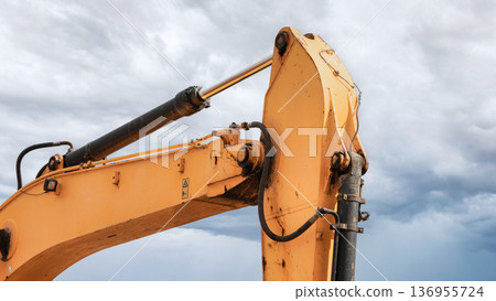 A robust yellow digger stands ready for action beneath a dramatic sky filled with swirling clouds, showcasing its strength and precision in construction 136955724