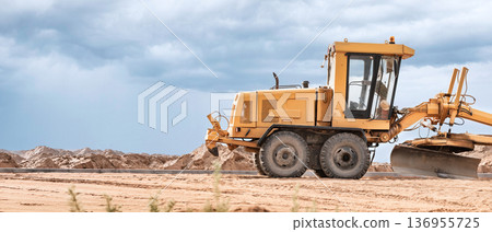 A yellow grader is actively leveling the sandy terrain at a construction site while dramatic clouds loom in the sky during late afternoon 136955725