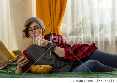 Caucasian teenage woman student resting indoors during free time. Girl eats popcorn and using smartphone. Home comfort theme. Mid shot Caucasian teenage woman student resting indoors during free time. Girl eats popcorn and using smartphone. Home comfort theme. Mid shot 136957511