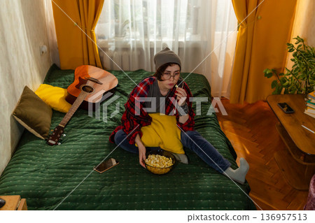 Young Caucasian teenage woman spending free time indoors. She watches TV and eats popcorn on her bed. Home relaxation theme. Top view, wide shot 136957513