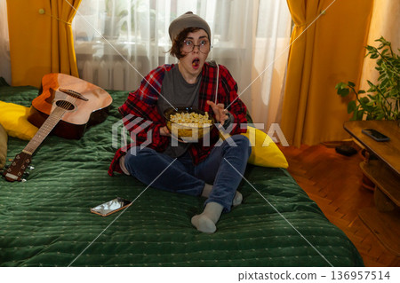Shocked Caucasian female teenager enjoying television at home. She sits on bed in casual clothes and eats popcorn. Relaxation theme. 136957514