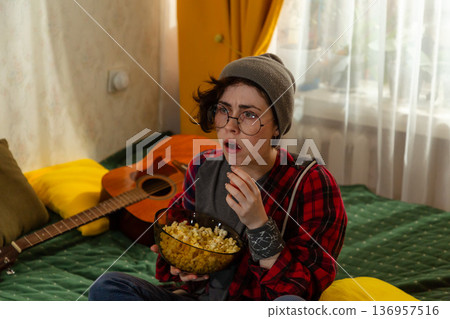 Portrait of Young Caucasian teenage woman enjoying television indoors. She relaxes on bed and eats popcorn. Home lifestyle concept. Portrait of Young Caucasian teenage woman enjoying television indoors. She relaxes on bed and eats popcorn. Home lifestyle concept. 136957516