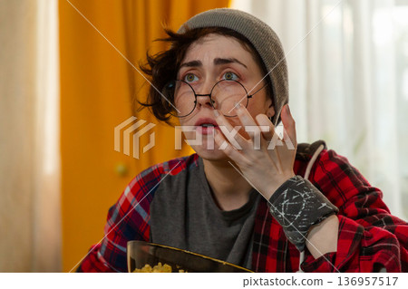 Portrait of young amazed Caucasian teenage woman relaxing after study time. She watches TV on bed and eats popcorn. Student lifestyle concept. 136957517