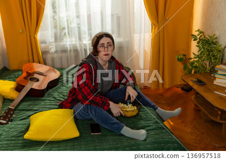 Caucasian teenage woman student enjoying a calm evening indoors. She watches television on bed and eat popcorn. Home comfort theme. Top view Caucasian teenage woman student enjoying a calm evening indoors. She watches television on bed and eat popcorn. Home comfort theme. Top view 136957518