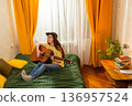 Dreaming Caucasian woman student practicing guitar in bedroom at home. She sits comfortably on a bed. Artistic skills development concept. Wide shot 136957524