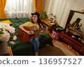 Happy young woman plays guitar in a home setting developing musical skills and creativity surrounded by plants and calm domestic atmosphere. 136957527