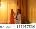 Young Caucasian woman folding clothes in bedroom at sunset time. She arranges personal items. Home care theme. Mid shot 136957530