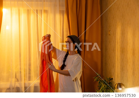Young Caucasian woman folding clothes in bedroom at sunset time. She arranges personal items. Home care theme. Mid shot Young Caucasian woman folding clothes in bedroom at sunset time. She arranges personal items. Home care theme. Mid shot 136957530
