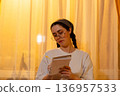Attractive young woman wearing eyeglasses keeping a journal at home during golden hour. Calm evening routine and mindfulness practice. Concept of mental health, and productivity in modern lifestyle. 136957533