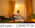 Young Caucasian woman folding clothes indoors during calm sunset hours. She organizes her living space. Home cleaning concept. Low angle view, wide shot 136957536