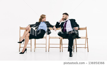 Stylish man and woman seated on wooden chairs facing each other in minimal white studio 136958366