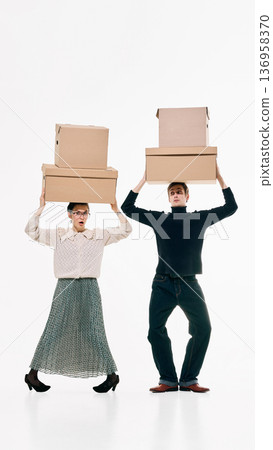 Stylish man and woman balancing stacked cardboard boxes on heads against white background. Stylish man and woman balancing stacked cardboard boxes on heads against white background. 136958370