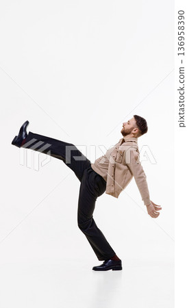 Stylish man in beige jacket and dark trousers performing high kick pose on white studio background. Stylish man in beige jacket and dark trousers performing high kick pose on white studio background. 136958390