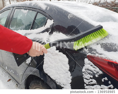 Person brushing fresh snow off black car with green snow brush on cold winter day. Person brushing fresh snow off black car with green snow brush on cold winter day. 136958985