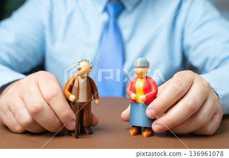 A man holding figurines of pensioners. An official and elderly people of retirement age. Social security and benefits administration. Government support for seniors A man holding figurines of pensioners. An official and elderly people of retirement age. Social security and benefits administration. Government support for seniors 136961078