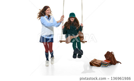 Teen girl pushing friend on swing with school bags isolated on white. 136961986