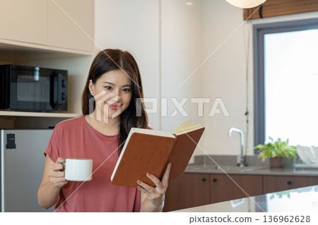 Smiling Asian Woman Reading Book with Coffee in Kitchen Smiling Asian Woman Reading Book with Coffee in Kitchen 136962628