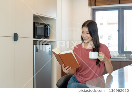 Smiling Asian Woman Reading Book with Coffee in Kitchen Smiling Asian Woman Reading Book with Coffee in Kitchen 136962630