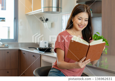 Smiling Asian Woman Reading Book with Coffee in Kitchen 136962631
