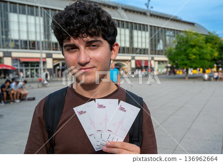Berlin, germany, august 15, 2023. Young man with curly hair smiling and holding berlin tv tower tickets at alexanderplatz, ready for sightseeing and city adventure 136962664