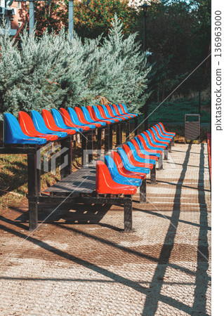 Colorful stadium seats lined up in a park during the day with greenery in the background Colorful stadium seats lined up in a park during the day with greenery in the background 136963000
