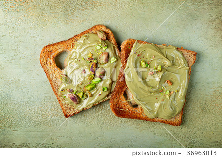 Two slices of toasted bread, topped with pistachio paste and sprinkled with pistachios, spread on a light surface 136963013