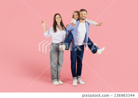 Playful father carrying daughter on shoulders beside smiling mother on pink background 136963344