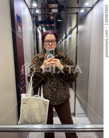 Woman taking mirror selfie in elevator wearing leopard coat. Urban lifestyle, personal style and daily commute routine. 136963905
