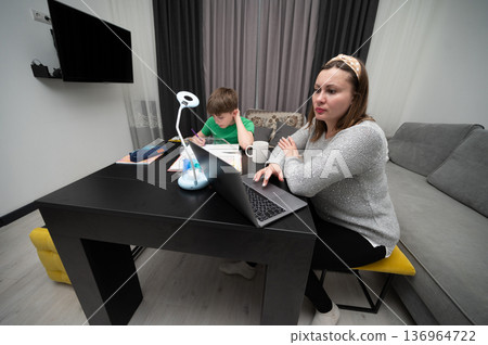 In a modern living room bathed in afternoon light, a mother diligently works on her laptop while her child focuses on their studies at home, creating a peaceful atmosphere 136964722