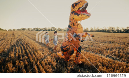 Inflatable Dinosaur T-Rex Is Chased, Running From Little Kids Boys In Field.  136964859